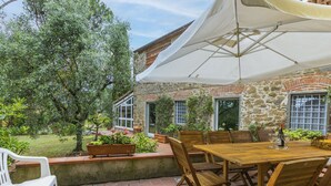 Een terras met een houten tafel en stoelen, een witte parasol en een stenen gebouw op de achtergrond.