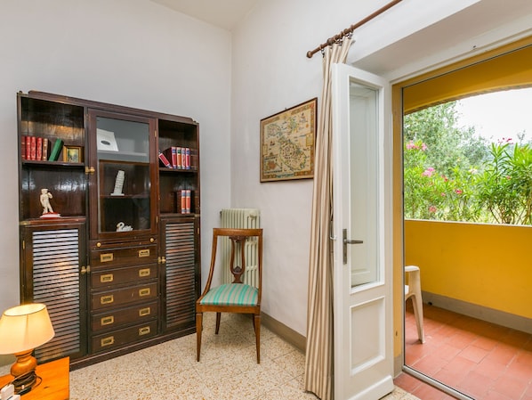 Una habitación con un armario de madera, una silla, un radiador, una puerta que da a un balcón y una pequeña mesa con una lámpara.