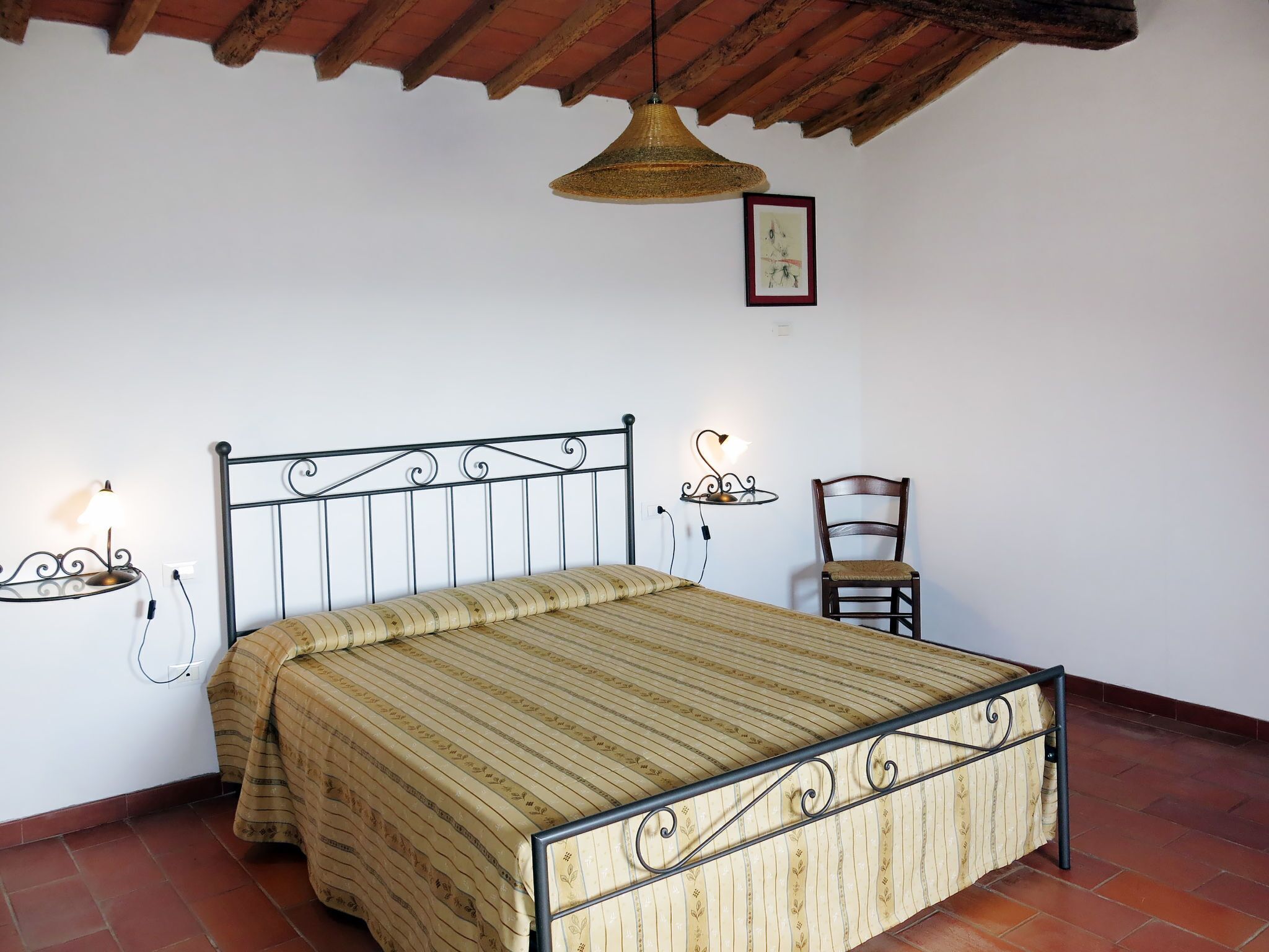 Una camera da letto con una struttura del letto in metallo, un comodino con una lampada, una sedia e una lampada a sospensione.