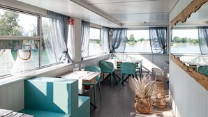A dining area with tables and chairs, large windows with curtains, and a view of the water outside.