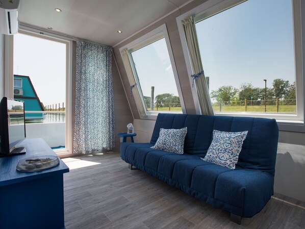 A modern room with a blue sofa, a small table with a glass bowl, and large windows offering a view of a blue building and greenery.