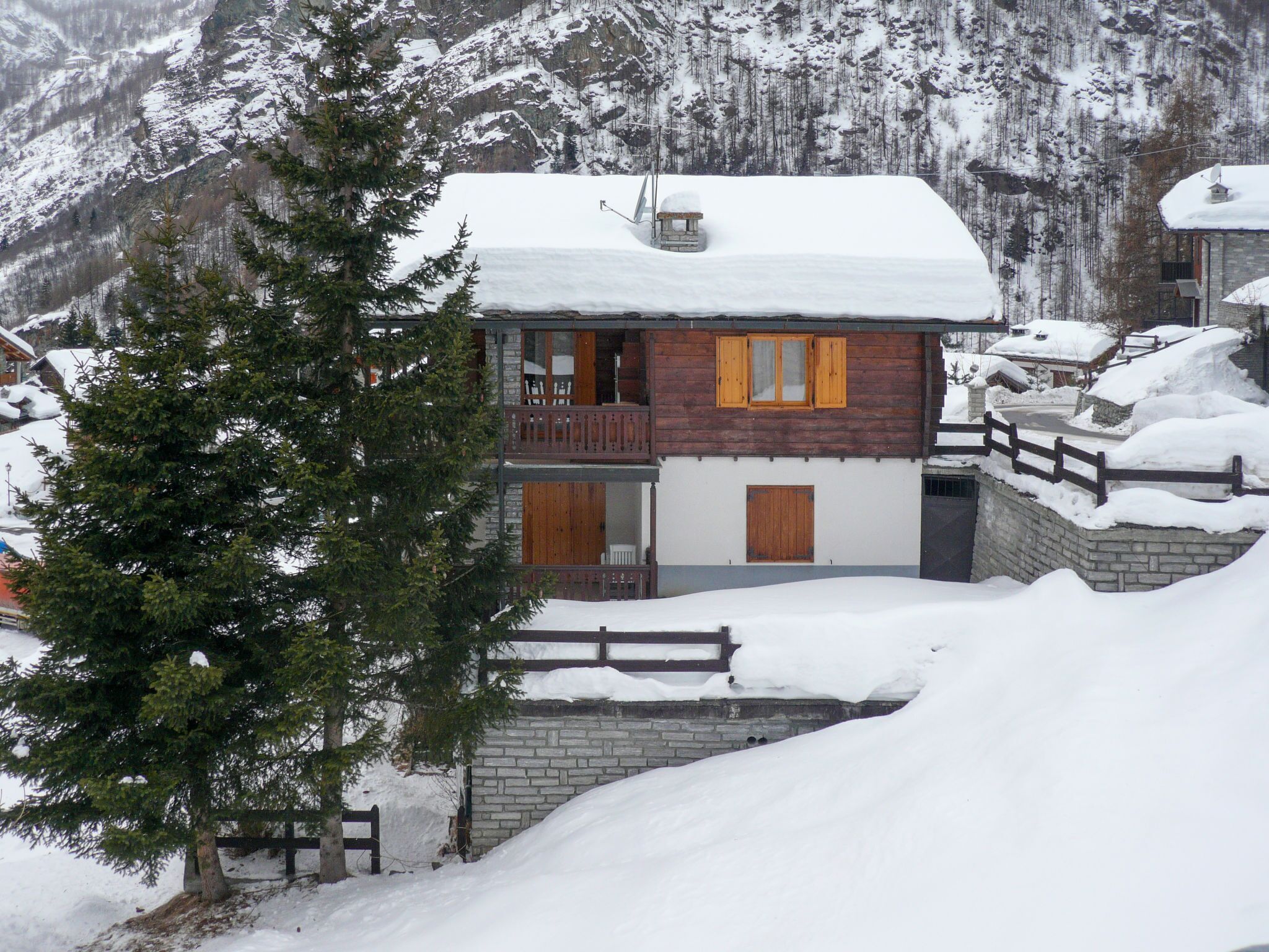 Eine zweistöckige Holzhütte mit Balkon, umgeben von schneebedeckten Bergen und Bäumen.