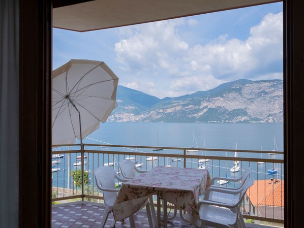 Un balcon avec une table et des chaises, un parasol, et une vue sur une étendue d’eau où l’on aperçoit des bateaux et des montagnes en arrière-plan.
