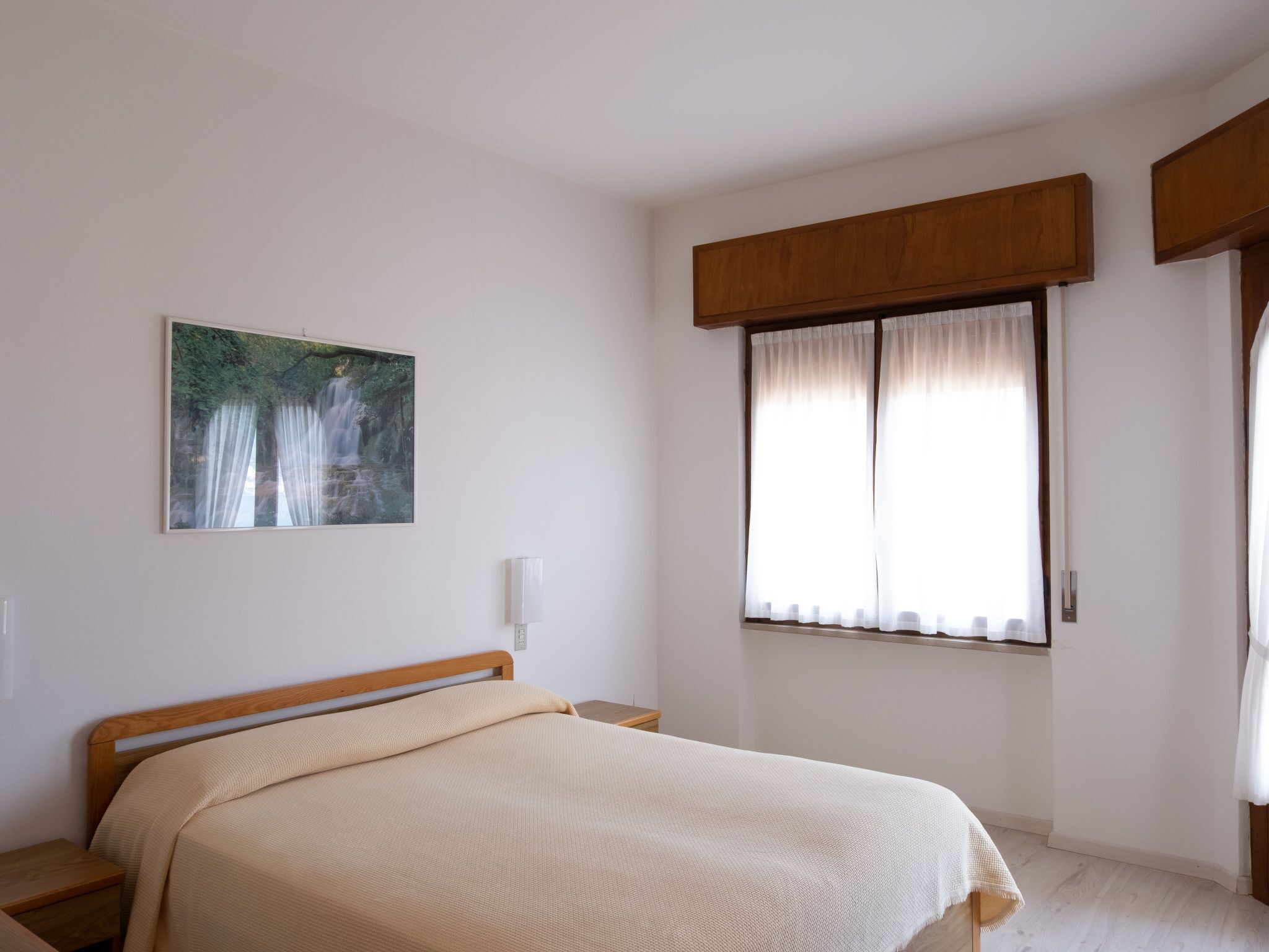 Un dormitorio con cama, mesita de noche, un cuadro y una ventana con cortinas.