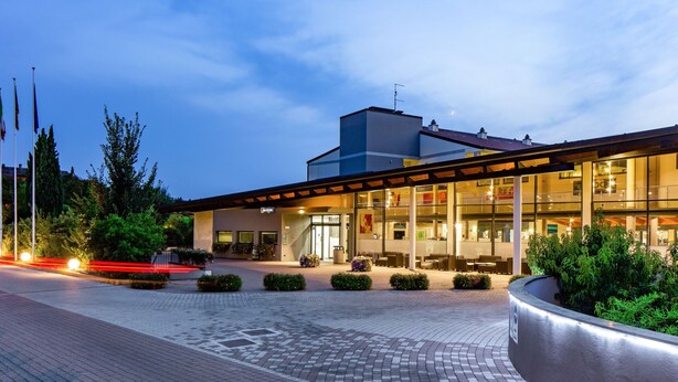 A modern hotel with a glass facade, a covered outdoor seating area, and a landscaped courtyard. - Apartment Eden by Interhome (Peschiera del Garda)