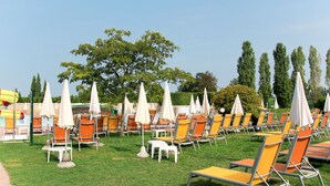 A poolside area with rows of orange and white lounge chairs and umbrellas, a grassy lawn, and a large tree in the background. - Apartment Eden by Interhome (Peschiera del Garda)