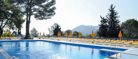 A swimming pool with sun loungers and umbrellas, surrounded by trees and mountains in the background.