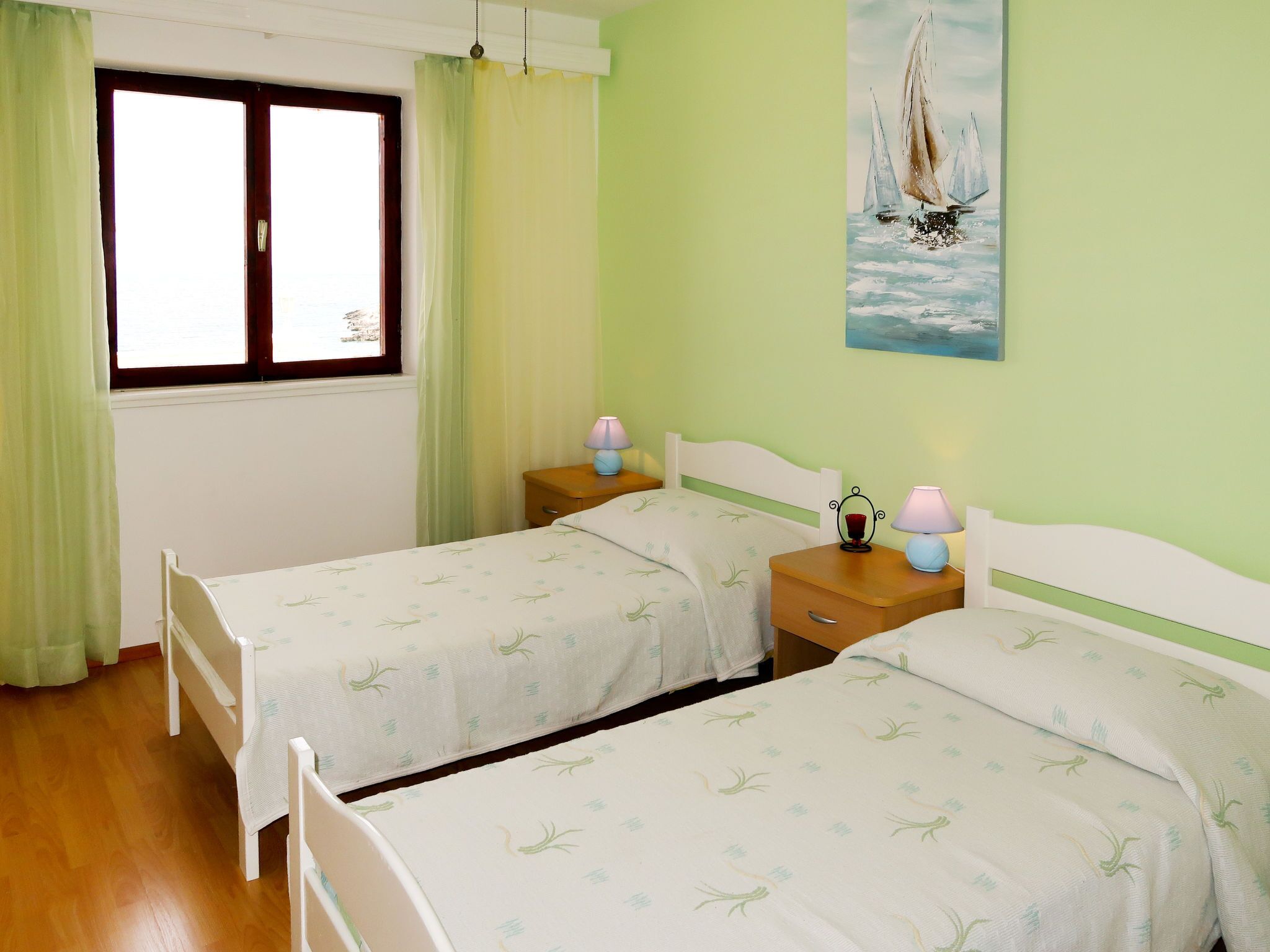 Un dormitorio con dos camas individuales, una ventana con cortinas, un cuadro de un velero y suelo de madera.