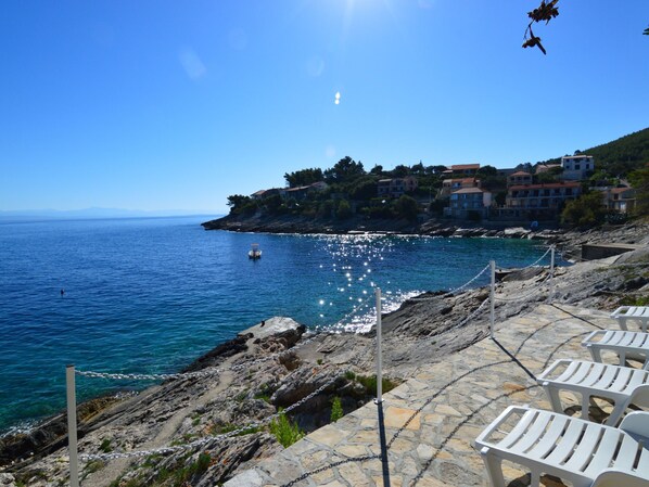 A coastal area with a rocky shoreline, a clear blue sea, and a row of white plastic chairs.