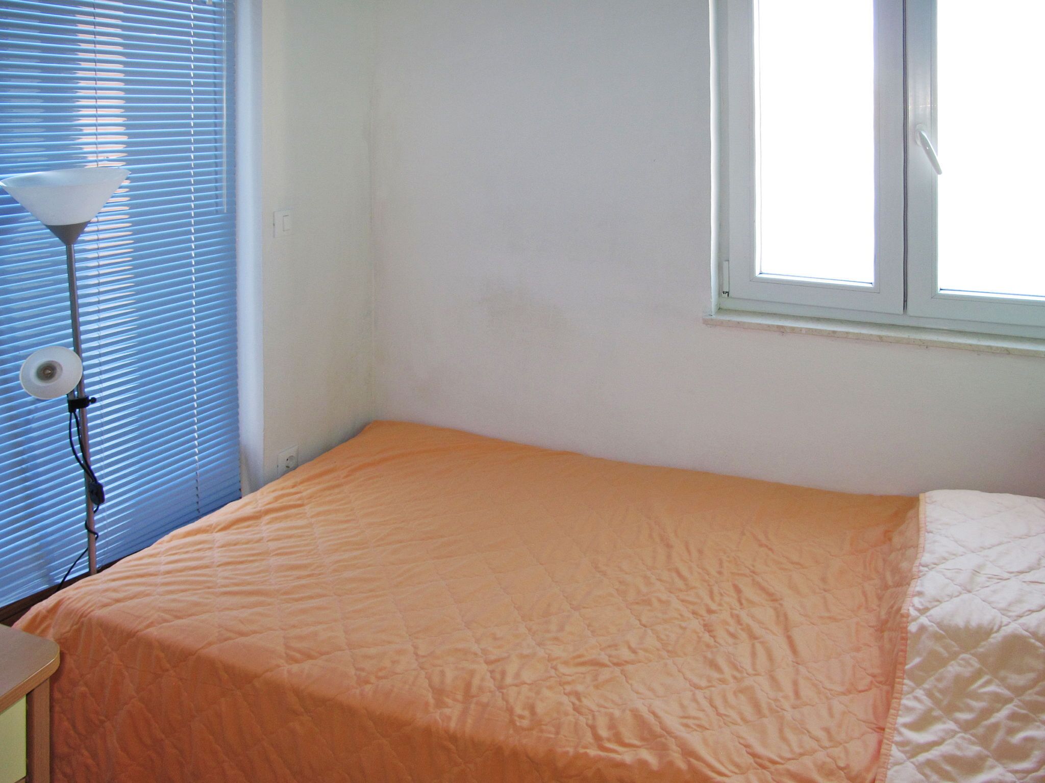 Une petite chambre avec un lit simple, un mur blanc uni, une fenêtre avec des stores et une lampe de chevet.