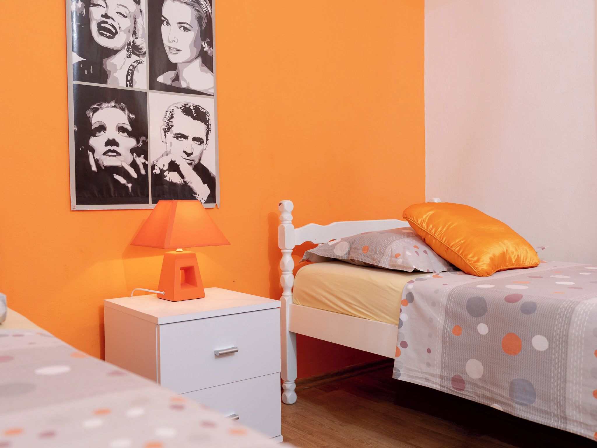 Een slaapkamer met een oranje wand, een witte nachtkastje met een lamp, een bed met een stippenbedekking, en een poster van een vrouw.