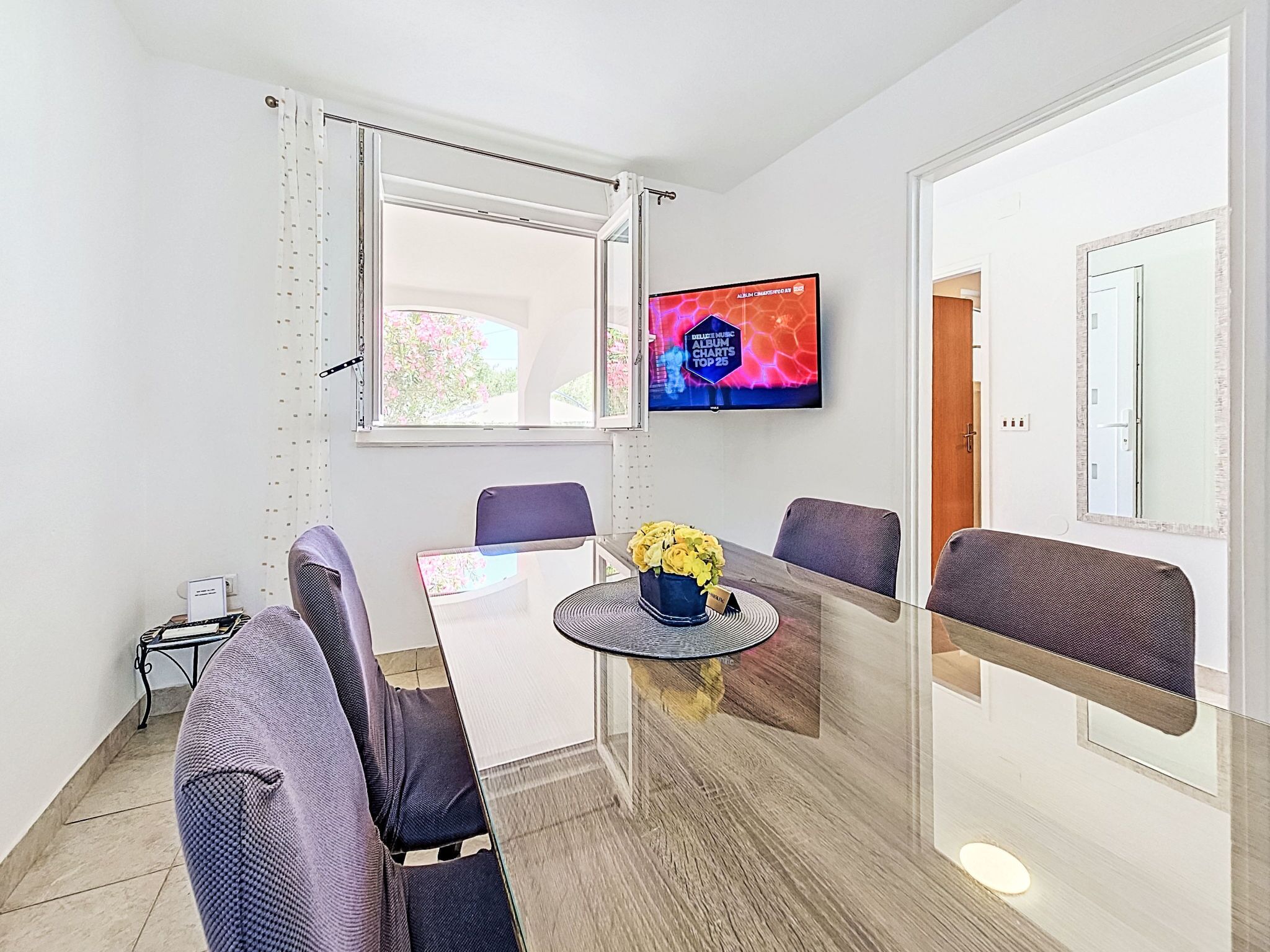 A modern room with a glass table, two chairs, a TV on the wall, and a window with curtains.