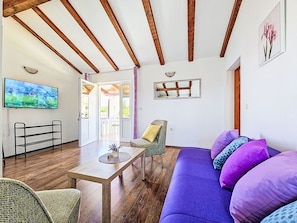 Een kamer met een houten plafond, een bank, een tafel, een stoel, een televisie en een deur naar buiten.