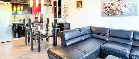 Een moderne woonkamer met een zwarte leren hoekbank, een glazen eettafel met stoelen, en een keuken op de achtergrond.