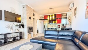 A modern living room with a sectional sofa, a flat-screen TV, a fireplace, and a kitchen area with red cabinets and pendant lights.
