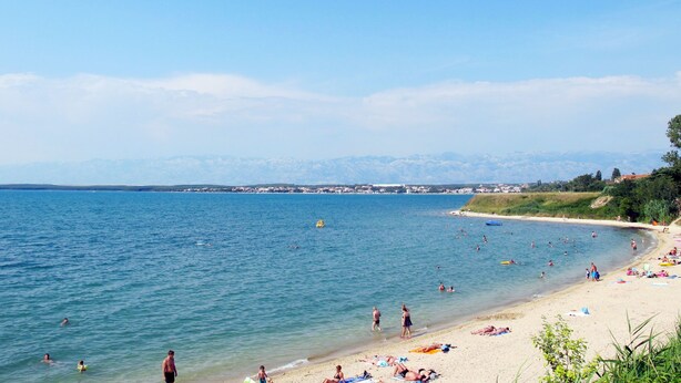 A beach with people swimming and sunbathing, a clear sky, and mountains in the background.