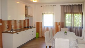 Een moderne keuken met witte kasten, een gasfornuis en een spoelbak. Er is een eethoek met een tafel en stoelen, en een raam met gordijnen.