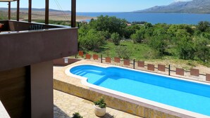 En swimmingpool med en terrass av sten, omgiven av grönska och med utsikt över havet och bergen.