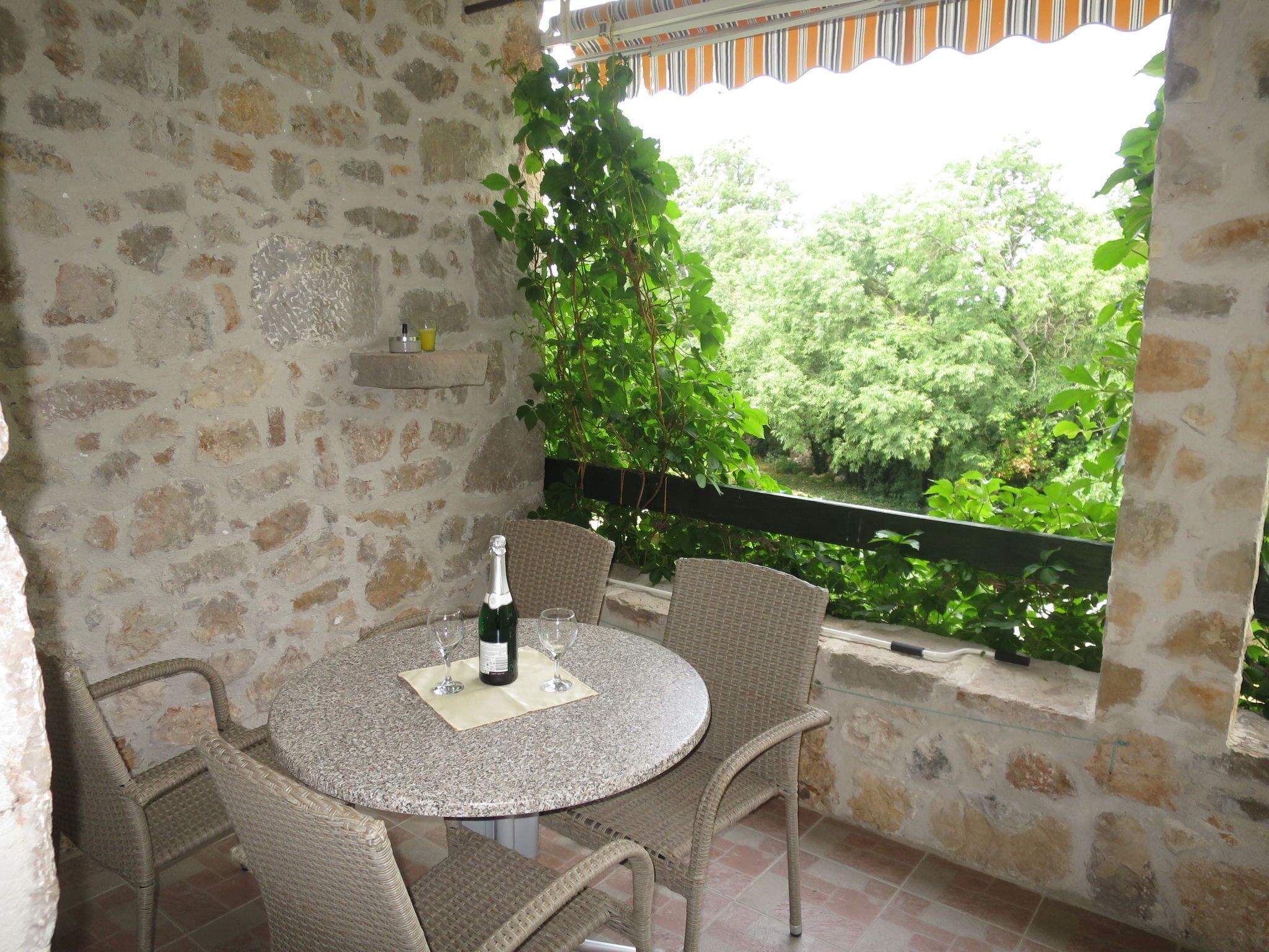 A stone wall, a round table with chairs, a bottle of wine, and a view of greenery.
