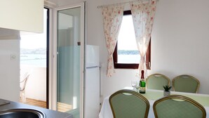 Una zona de menjador amb una taula, cadires, una ampolla de vi i vista al mar a través d'una finestra amb cortines florals.