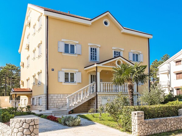 A two-story building with a yellow facade, multiple windows with shutters, a central entrance with a staircase, and a stone wall surrounding the property.