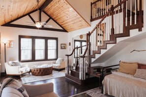 Un salon spacieux avec un plafond haut, un escalier en bois et un coin salon confortable agrémenté d’un pouf rond.