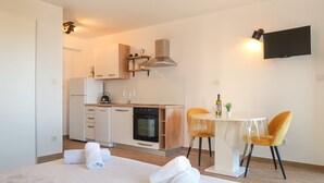 Een moderne hotelkamer met een bed, een kitchenette met fornuis en spoelbak, een eettafel met stoelen, en een flatscreen televisie aan de wand.