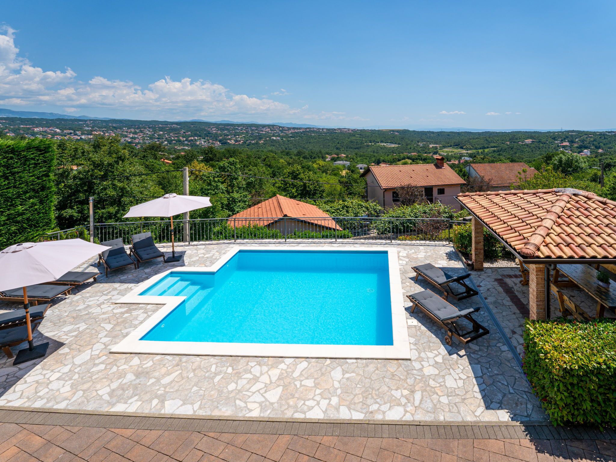 A swimming pool with stone tiles, surrounded by lounge chairs and umbrellas, with a view of houses and greenery in the background.