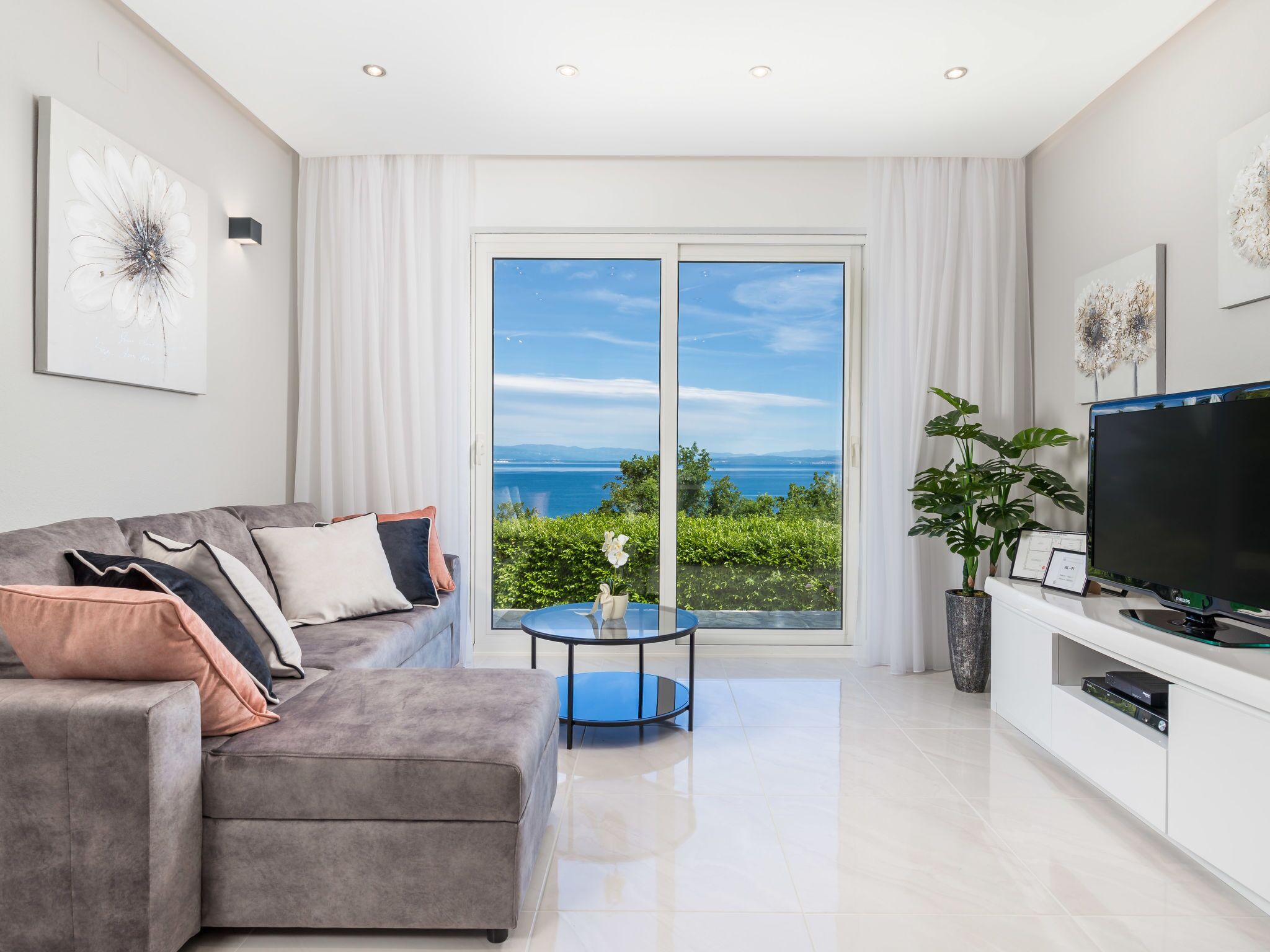 Uma sala de estar moderna com um sofá cinzento, uma televisão de tela plana e uma janela grande com vista para o mar.