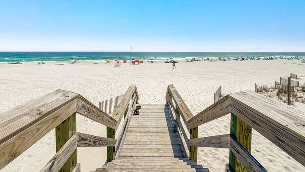 Un escalier en bois mène à une plage de sable fin, sous un ciel dégagé.