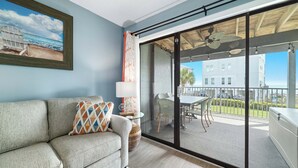 Un salon moderne avec un canapé, une porte vitrée donnant sur une terrasse extérieure et un tableau représentant une scène de plage.