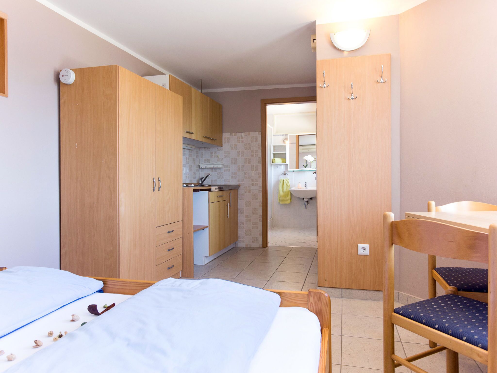 A compact room with a bed, a wooden chair, a small table, and a kitchenette area.