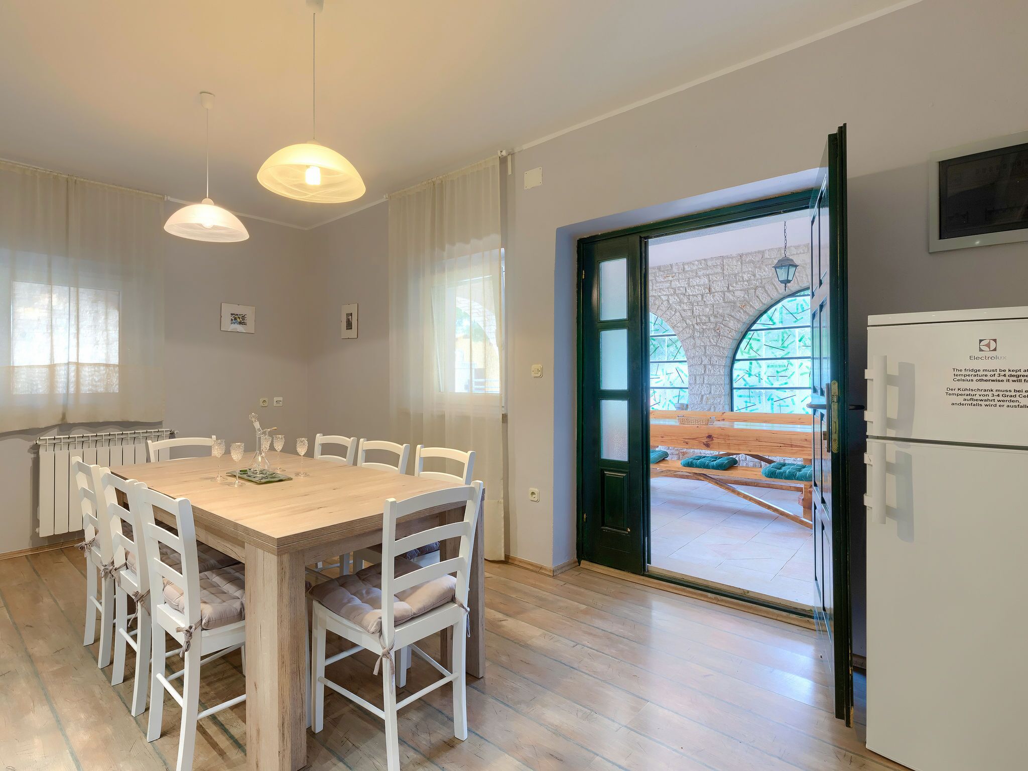 Un comedor con mesa de madera y sillas blancas, un refrigerador y vista a un área de estar exterior por una puerta de vidrio.