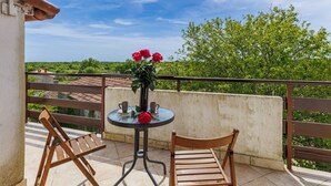 A balcony with a table and two chairs, a vase with red flowers, and a view of green trees and buildings. - Vacation home Lancin by Interhome (Pula/Marčana)