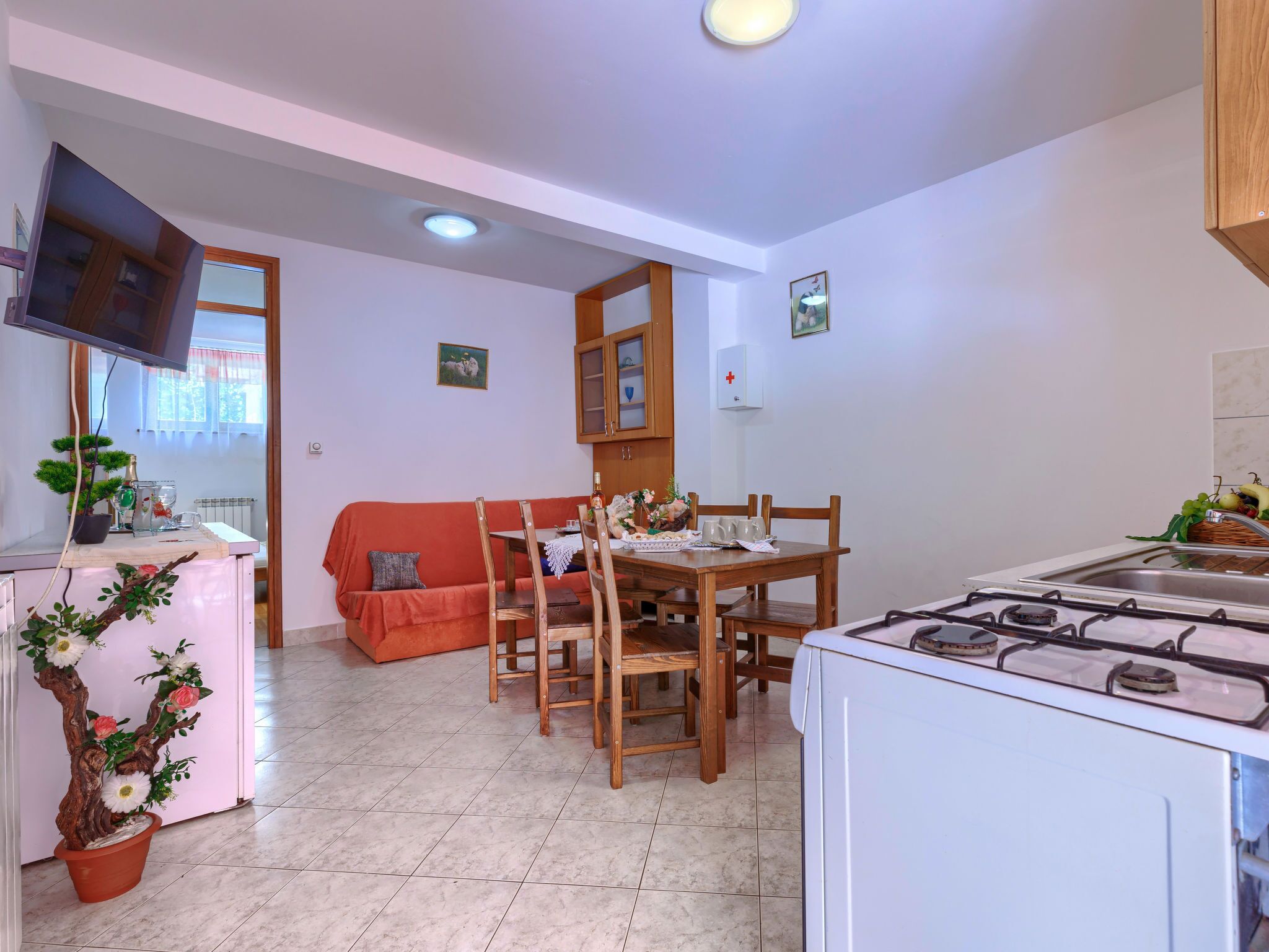 A compact kitchen with a gas stove, wooden dining table, and chairs. A small TV is mounted on the wall, and there is a potted plant near the entrance.