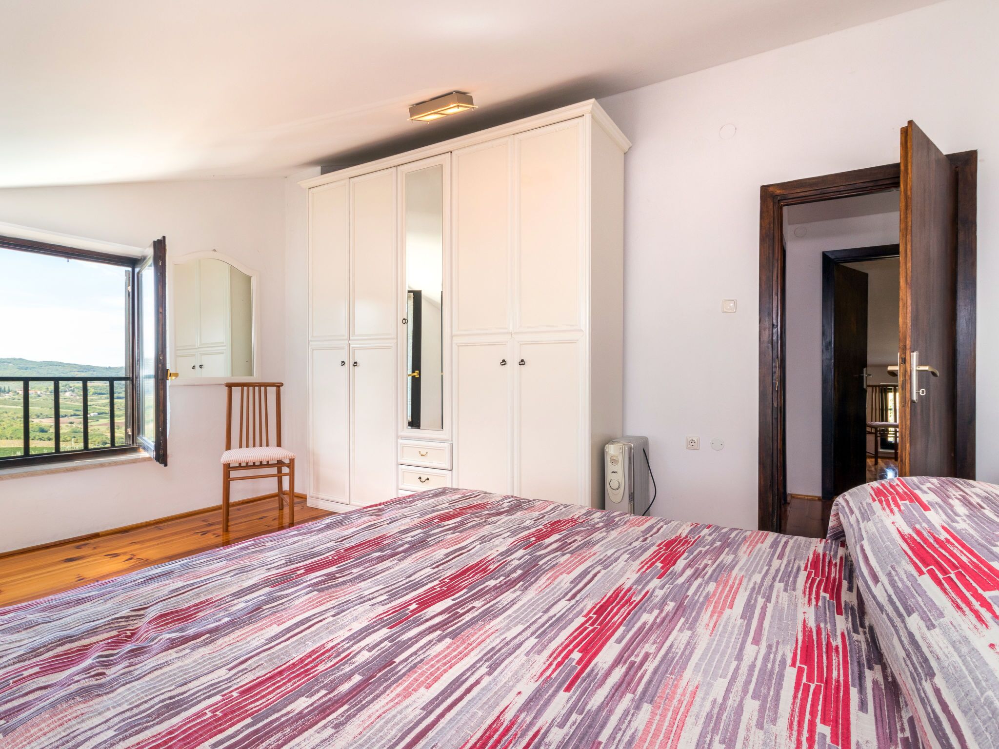 Ein Schlafzimmer mit großem Kleiderschrank, einem Bett mit gemusterten Textilien, einem Sessel und Blick durch ein Fenster.