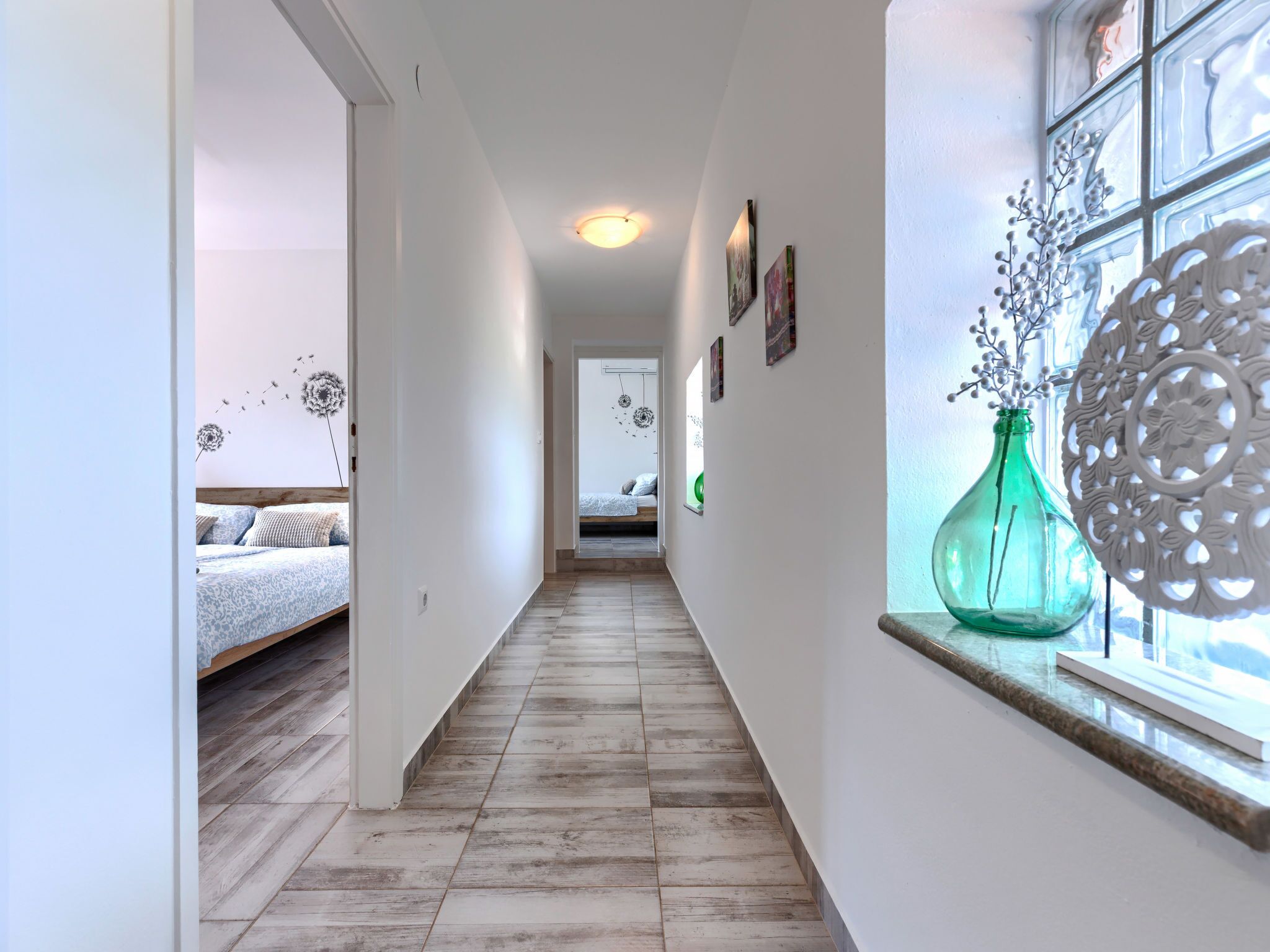 A narrow hallway with a wooden floor leading to a bedroom with a bed and a window with decorative items.