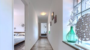 A narrow hallway with a wooden floor leading to a bedroom with a bed and a window with decorative items.