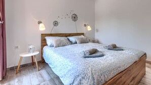 A bedroom with a wooden headboard, a patterned bedspread, two bedside lamps, a small bedside table, and a wall decoration of dandelions.
