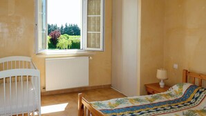 A bedroom with a crib, a bed with a floral bedspread, a window with a view of greenery, and a radiator. - Plan du Vas by Interhome (Valreas)