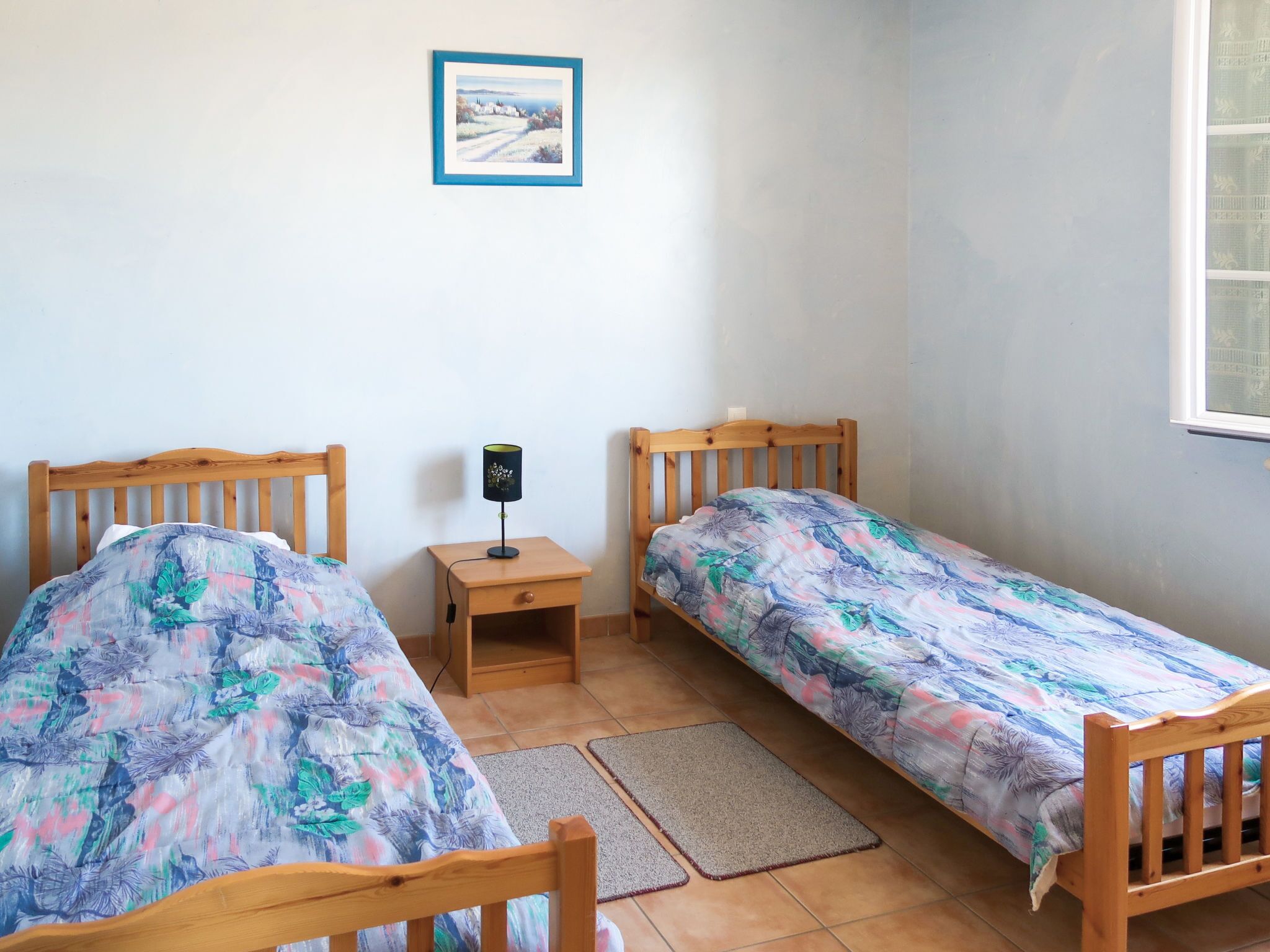 Une petite pièce propre et bien rangée, avec deux lits en bois, une table de chevet, une lampe et un tableau encadré au mur.