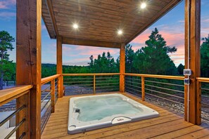 Eine Holzterrasse mit Whirlpool, umgeben von einem Geländer und Blick auf Bäume und einen Sonnenuntergang.