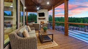 A wooden deck with wicker furniture, a fireplace, and a pool overlooking a forested area. - Featured on Staycation: The Overlook | Pool, Pickleball, ATV Access & Views (Broken Bow)