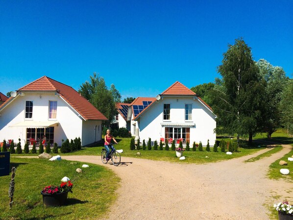 A row of white houses with red roofs, a cyclist on a dirt path, and a well-maintained garden with rocks and flowers.