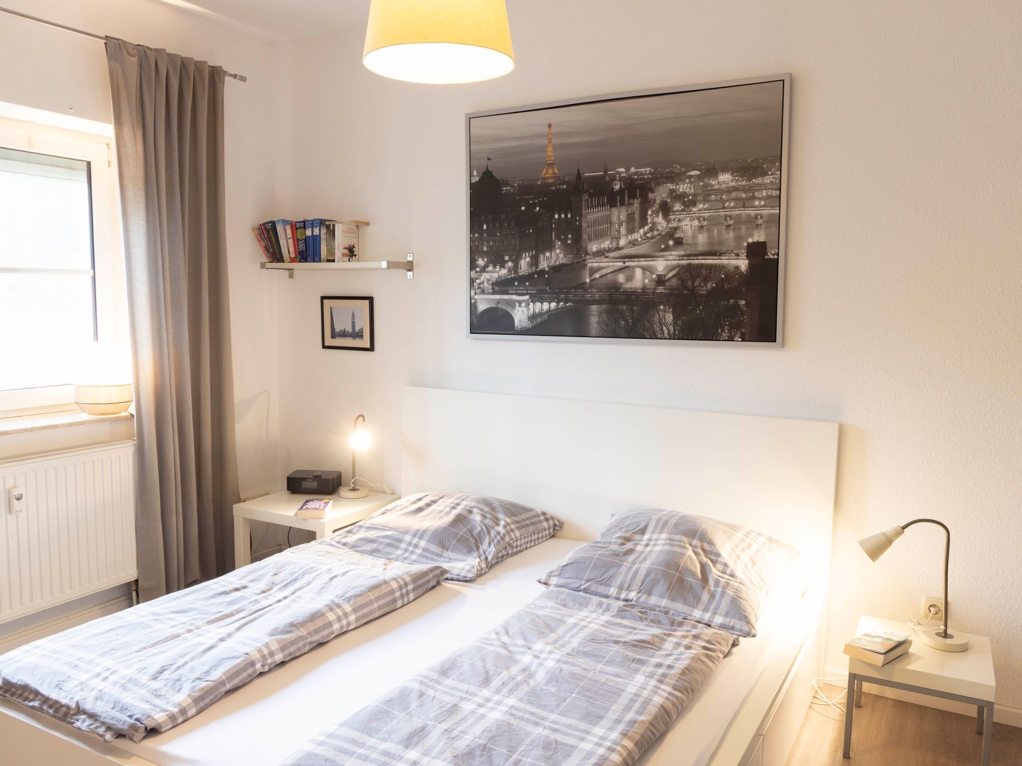 Une chambre à coucher avec un lit, des tables de chevet, une lampe, un tableau représentant un paysage urbain et une fenêtre avec des rideaux.