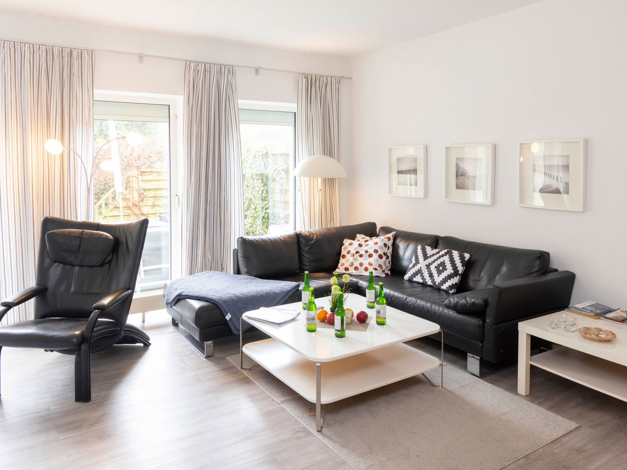 Una sala de estar moderna con un sofá y un sillón de cuero negro, una mesa de centro blanca con botellas y frutas, y una mesita auxiliar blanca con un plato.
