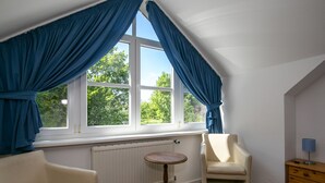 Une pièce avec un plafond en pente, un radiateur, une table ronde en bois, deux chaises, une commode et une fenêtre avec des rideaux bleus.