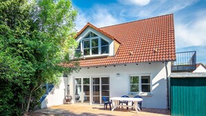 Ein zweigeschossiges Haus mit rotem Ziegeldach, weißen Außenwänden und einer überdachten Terrasse mit Tisch und Stühlen.