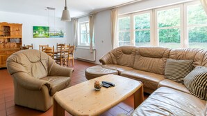 Ein Wohnzimmer mit einer Couch, einem Sessel und einem Couchtisch. Ein Esstisch mit Stühlen, ein Holzschrank und ein Gemälde an der Wand sind ebenfalls vorhanden. Große Fenster sorgen für natürliches Licht.