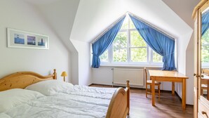 Ein Schlafzimmer mit Bett, Schreibtisch und Stuhl. Ein Fenster mit blauen Vorhängen und ein Bilderrahmen sind an der Wand.